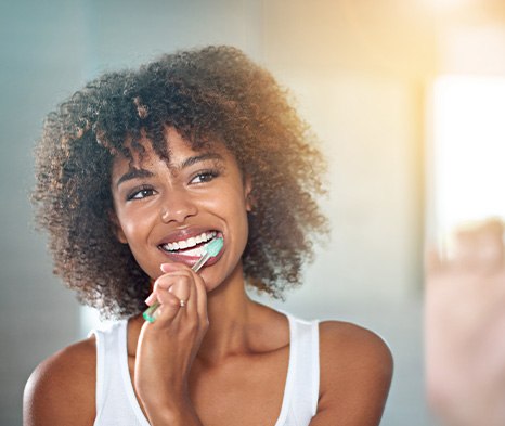 Woman in white tank top smiling brushing her teeth with glowing sunlight